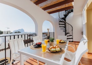 A dining table with breakfast items, white chairs, a spiral staircase, and a view of the city. - Casa Sunrise - Rooftop, 5 Swimming Pools and Panoramic Sea view @ Peñon de Ifach (Calp)