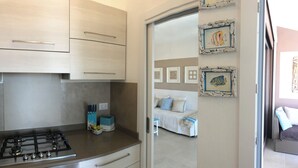 Une cuisine compacte équipée d’une cuisinière à gaz, d’un petit plan de travail et d’un placard. On y trouve également un coin repas avec une table et des chaises, et une chambre à coucher visible à travers une porte ouverte.