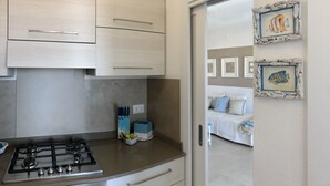 Une cuisine équipée d’une cuisinière à gaz, de placards et d’un plan de travail. On aperçoit un lit à travers une porte ouverte donnant sur la pièce adjacente.