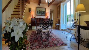 A spacious living area with a wooden staircase, a dining table with chairs, a chandelier, and a large window with curtains.