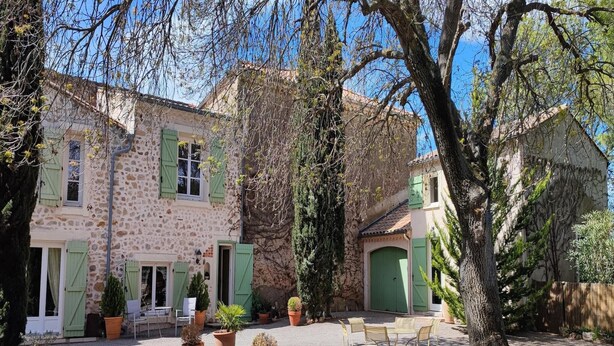 A stone building with green shutters, a courtyard with potted plants, and outdoor seating.