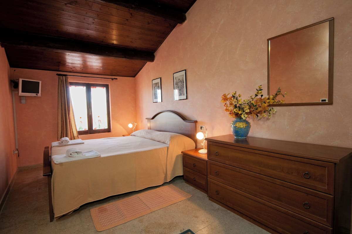 Ein Schlafzimmer mit Bett, Nachttisch, Kommode, Spiegel und einem Fenster mit Vorhängen.