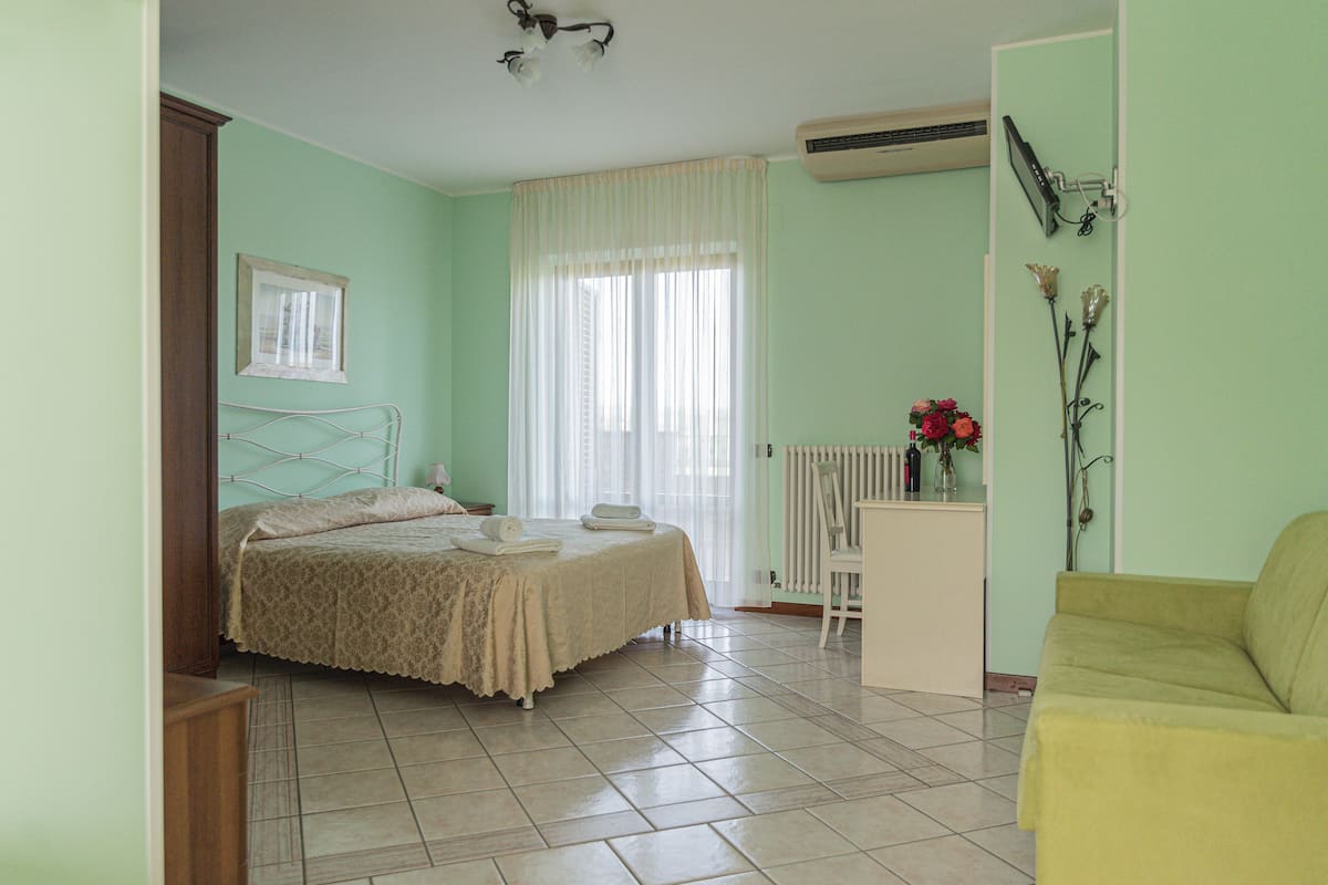 Ein Schlafzimmer mit einem Bett, einem Heizkörper, einem Fenster mit Vorhängen, einem kleinen Tisch und einem gelben Stuhl.