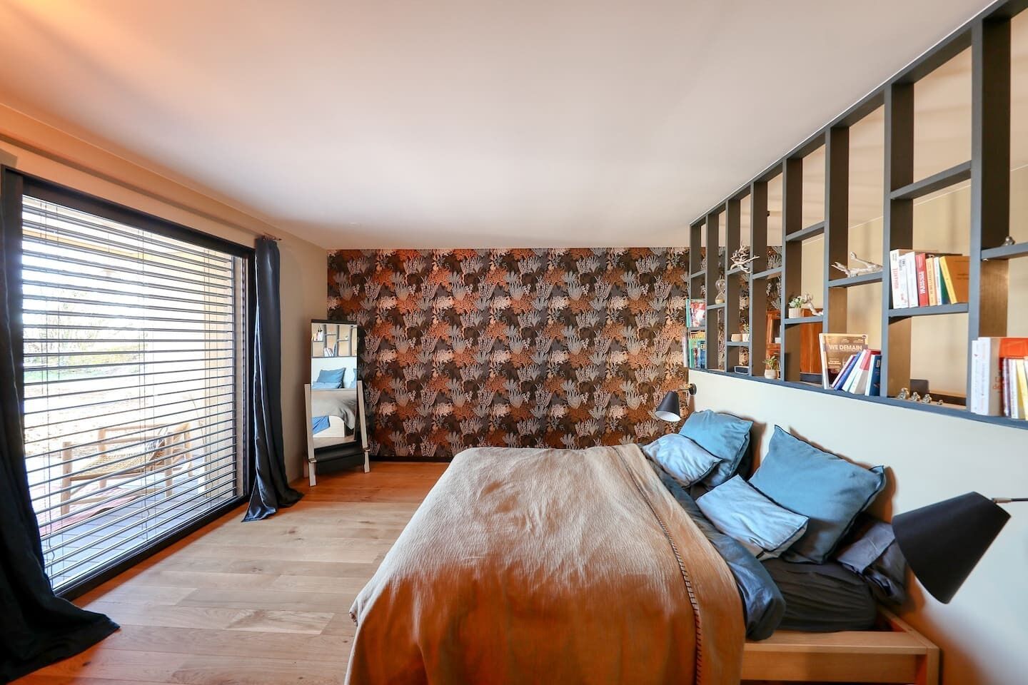 A bedroom with a large bed, a bookshelf, and a window with blinds.