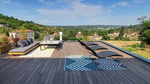 Une terrasse dotée d’un canapé, de deux fauteuils et d’une table, offrant une vue sur un quartier résidentiel avec ses maisons et ses collines.