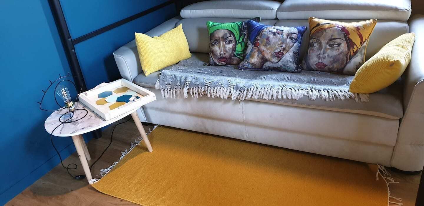 A living room with a grey sofa, a yellow throw pillow, a blue wall, and a small table with a lamp and a board game.