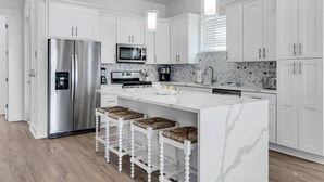Une cuisine moderne avec des meubles blancs, des appareils en acier inoxydable, un îlot en marbre et des luminaires suspendus.
