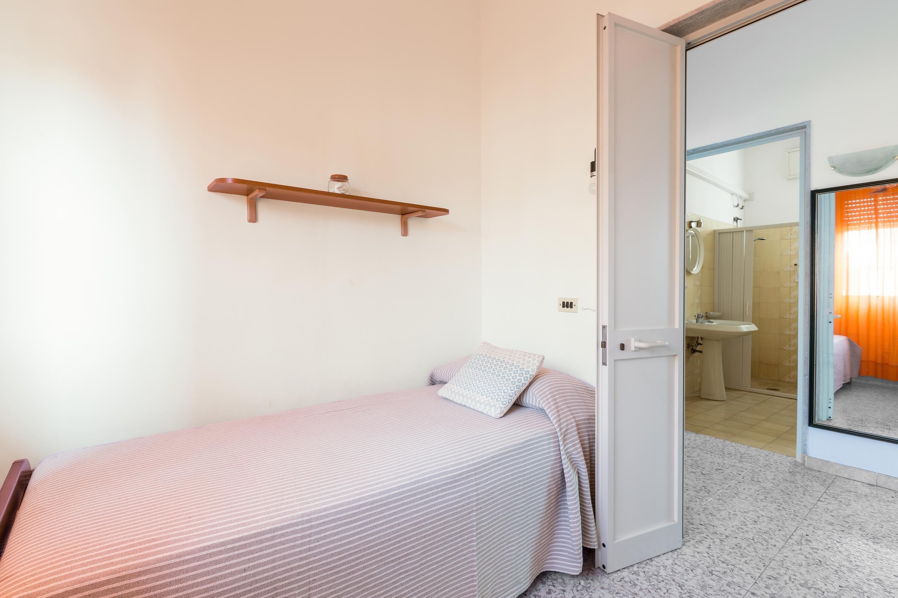 A bedroom with a single bed, a bedside table, and a small shelf. A bathroom is visible through an open door.