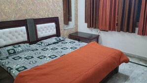 A bedroom with a bed, a bedside table, a mirror, and a window with curtains. - Roomshala 153 Hotel Krishnashrya (Mathura)