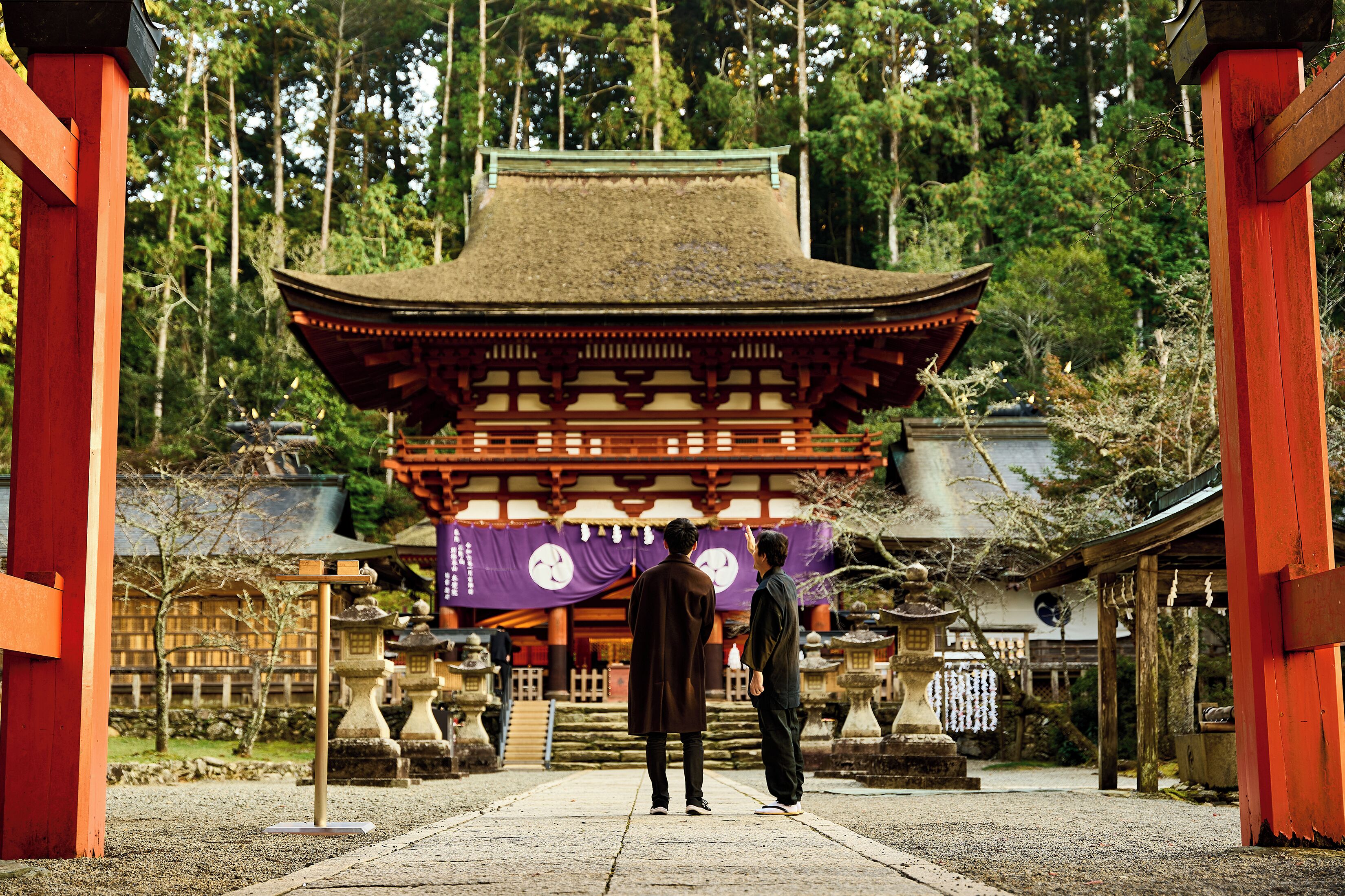 A traditional Japanese temple with a thatched roof, surrounded by trees and stone lanterns.