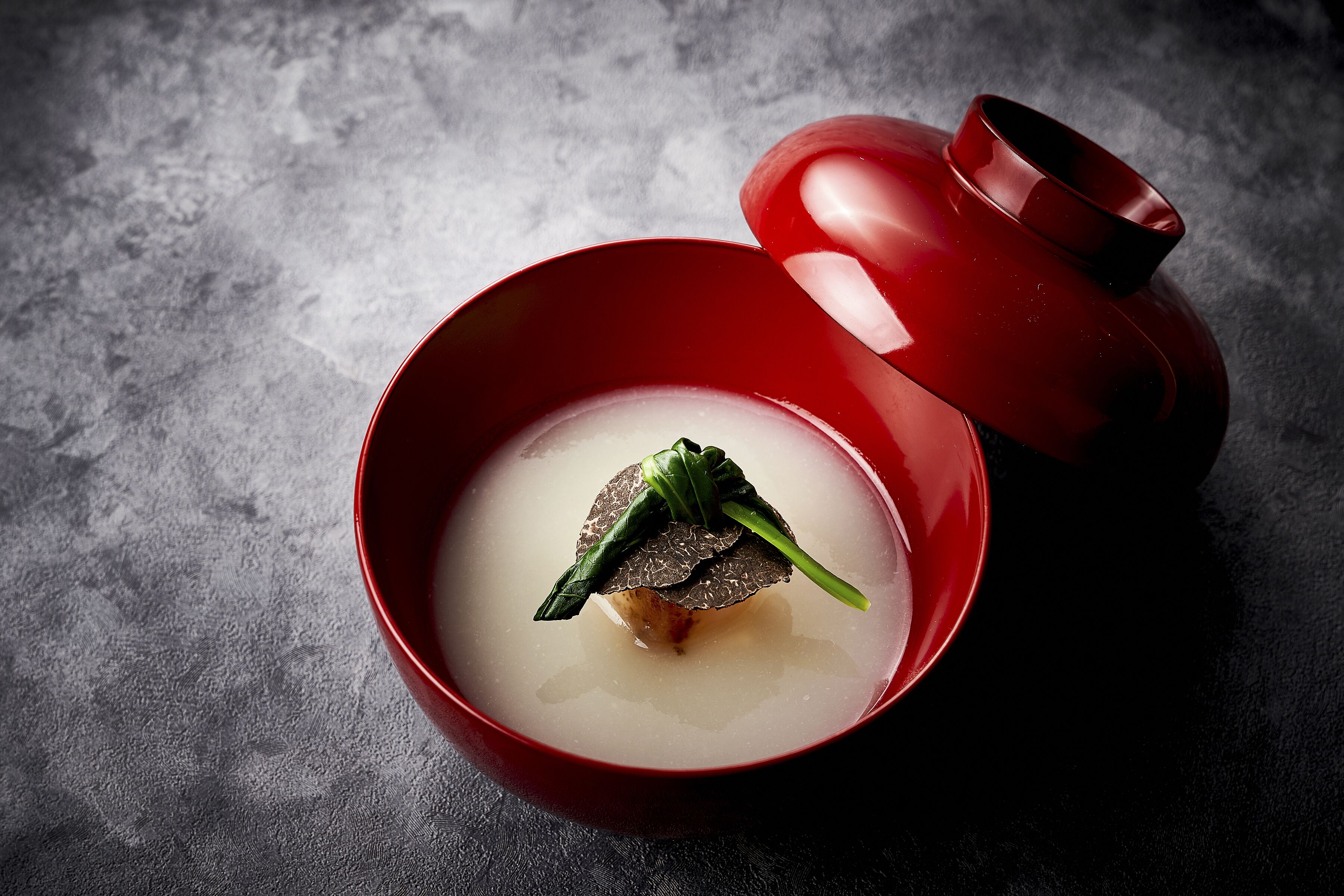 Una ciotola rossa con coperchio, contenente un liquido trasparente e un guarnizione di verdure verdi.