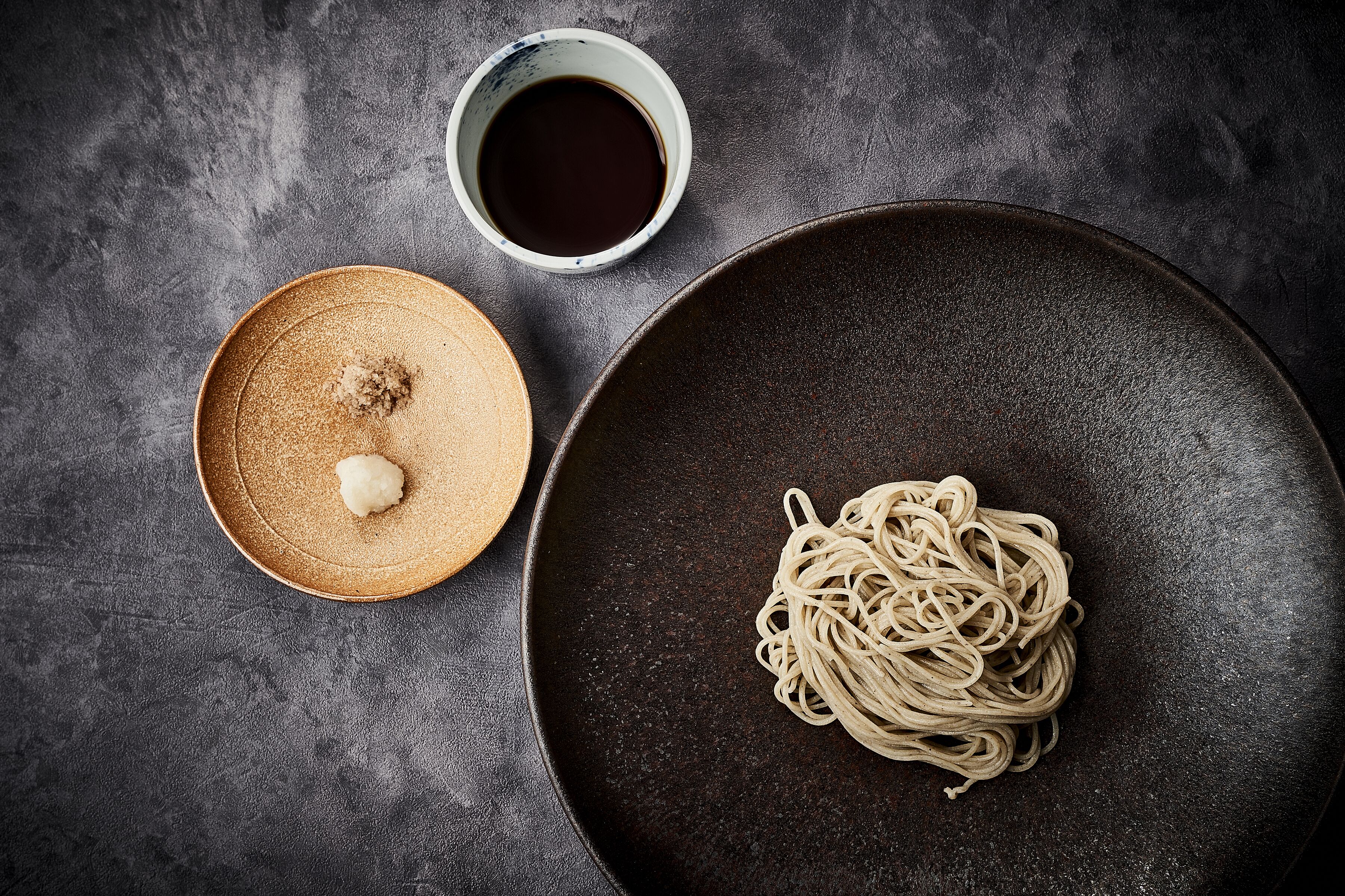 A dark textured surface with a bowl of noodles, a small bowl of sauce, and a plate with a dollop of sauce.