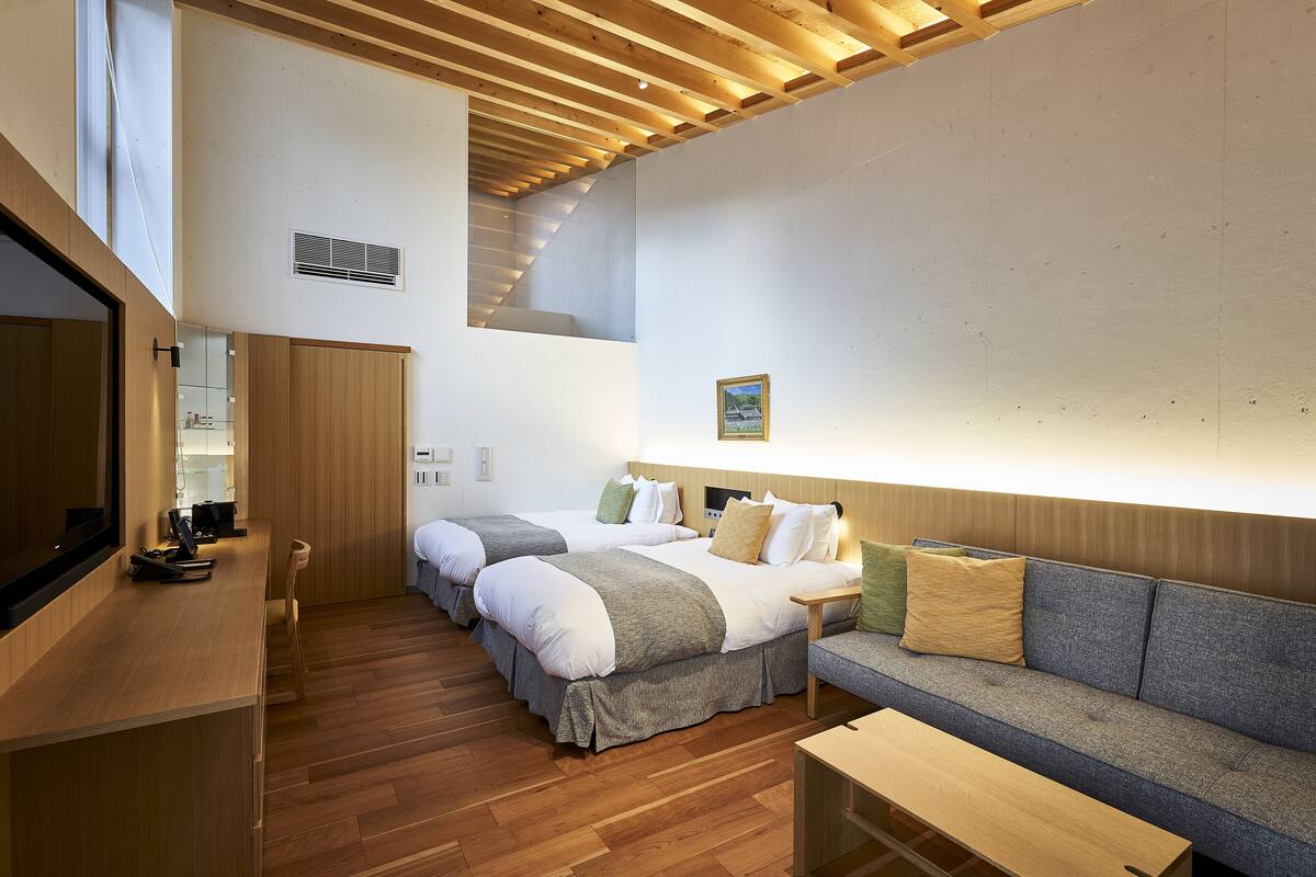 A modern hotel room with two beds, a sofa, a wooden desk, and a large mirror.