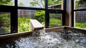 A hot tub with water flowing out, viewed through a window with a view of a wooden fence and greenery outside. - Sanso Amanosato (Katsuragi)