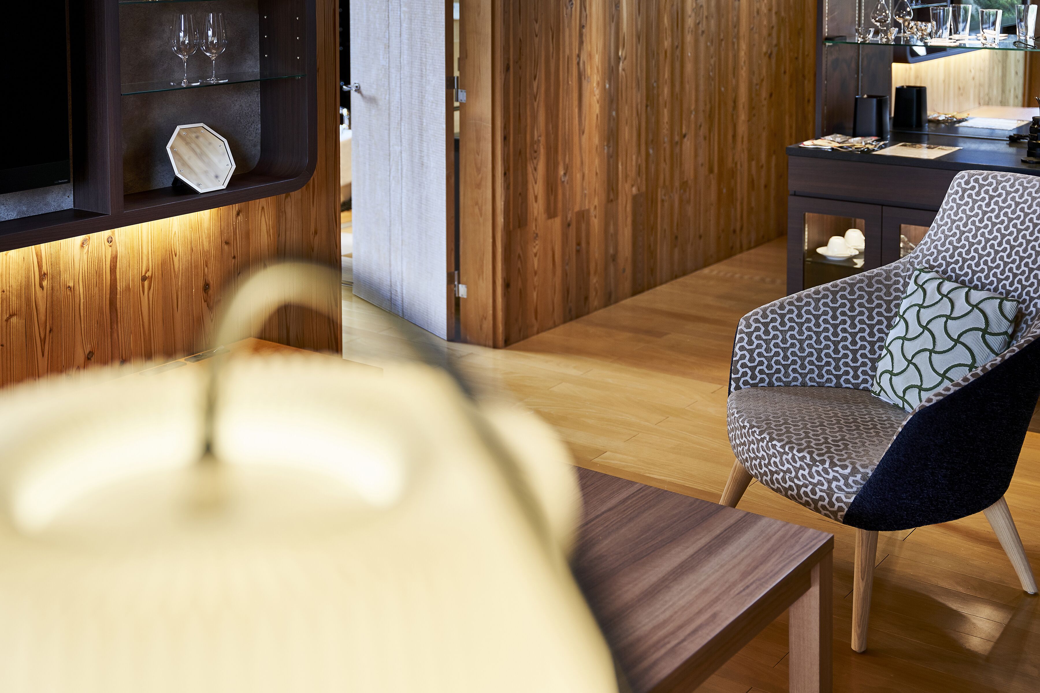 A modern interior with a wooden floor, a patterned armchair, a glass shelf with wine glasses, and a wooden cabinet.