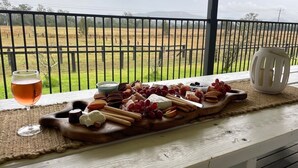 A wooden board with various cheeses, fruits, and crackers on a table by a window overlooking a field.