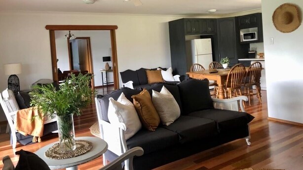 A living room with a black sofa, a dining area with wooden chairs, a kitchen with dark cabinets, and a ceiling fan.