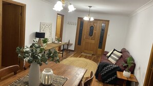 A dining area with a wooden table, chairs, a vase with plants, a small side table with a plant, and a sofa with a patterned throw.