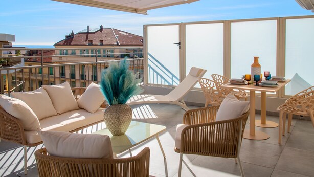 A sunlit balcony with wicker furniture, a glass table, and a view of buildings in the background. - Résidence Hôtelière Chambord (Menton)