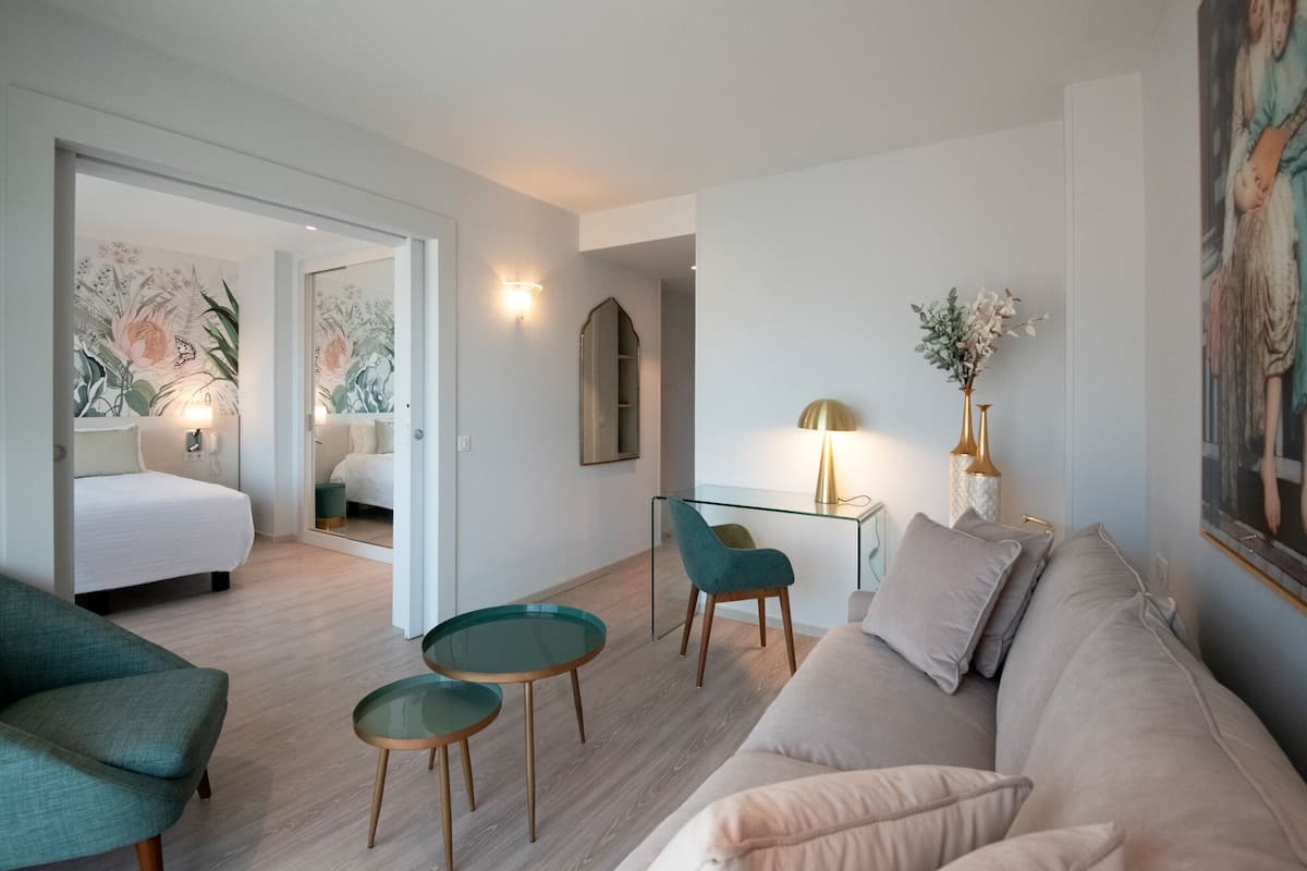 Ett modernt vardagsrum med en soffa, ett soffbord i glas, en turkos fåtölj och ett litet sidobord. I det intilliggande rummet syns en säng med en väggmålning med blommotiv.