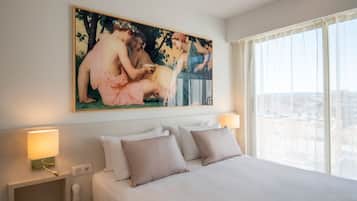 Ein Hotelzimmer mit einem großen Bett, zwei Nachttischlampen, einem Bild an der Wand und Blick auf die Stadt durch das Fenster.
