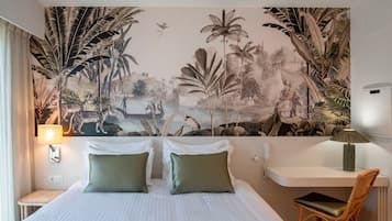 Hotelkamer met een bed, twee kussens, een nachtkastje, een bureau, een stoel en een groot muurdecoratie met een junglelandschap.