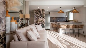 A modern living room with a sofa, a dining area with chairs, a kitchen with a refrigerator, and a large window. - Résidence Hôtelière Chambord (Menton)