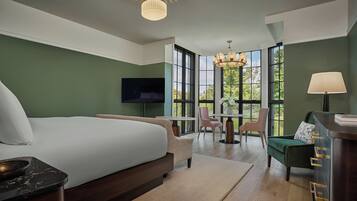 Une chambre d’hôtel moderne dotée d’un grand lit, d’une télévision, d’un coin repas avec des chaises et de grandes fenêtres offrant une vue sur l’extérieur.