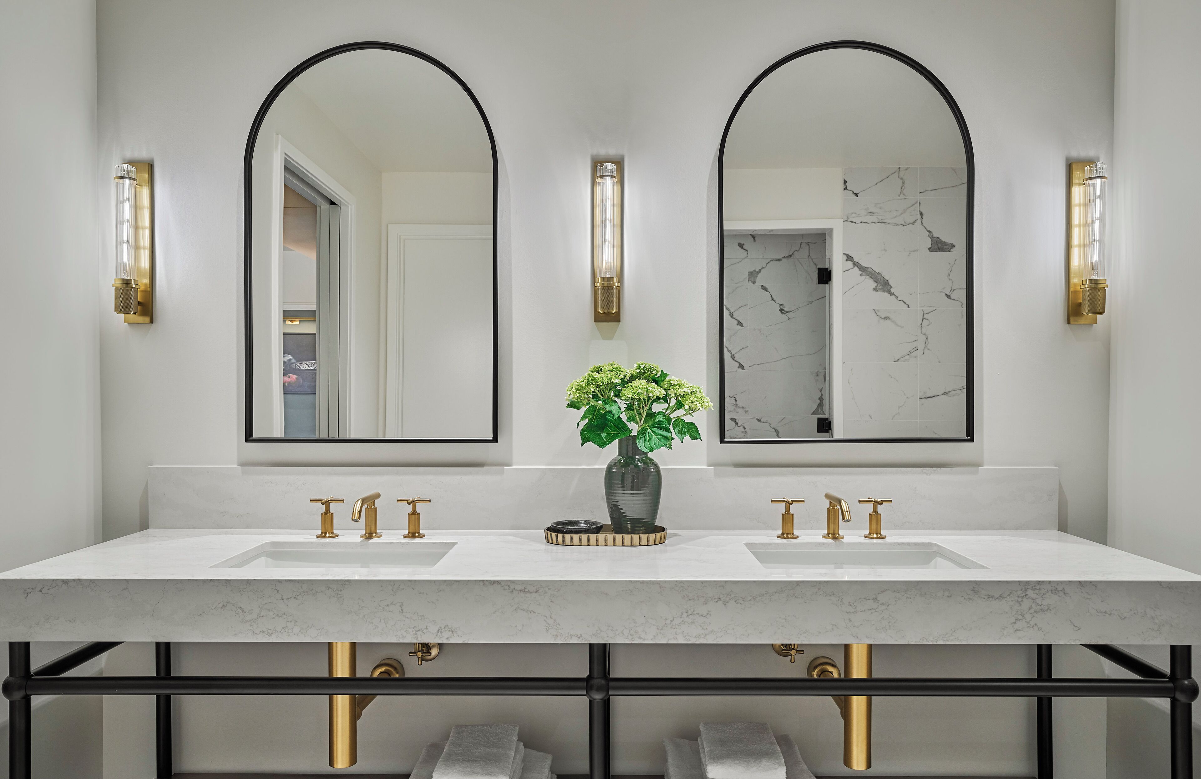 A bathroom with a white marble sink and vanity, two gold faucets, a vase with green plants, and two wall-mounted mirrors with black frames.