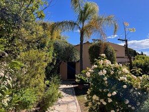 A garden with a stone pathway leading to a building with a dark entrance. - Les Petites Terrasses (Grasse)