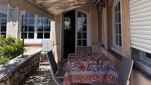 An outdoor patio with a table and chairs, a covered area, and a view of the house. - Les Petites Terrasses (Grasse)