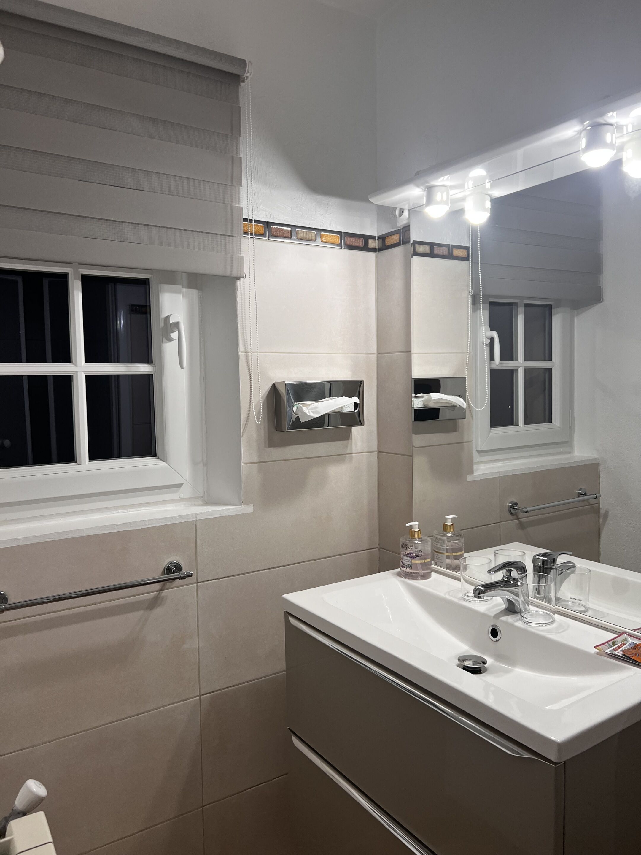 Un baño con lavamanos blanco, espejo, toallero y ventana con persianas.