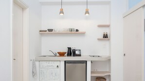 A compact kitchen with a dishwasher, a coffee maker, and a sink. There are wooden shelves above the counter and a basket below. - Flisvos Beach Apartments (Naxos)