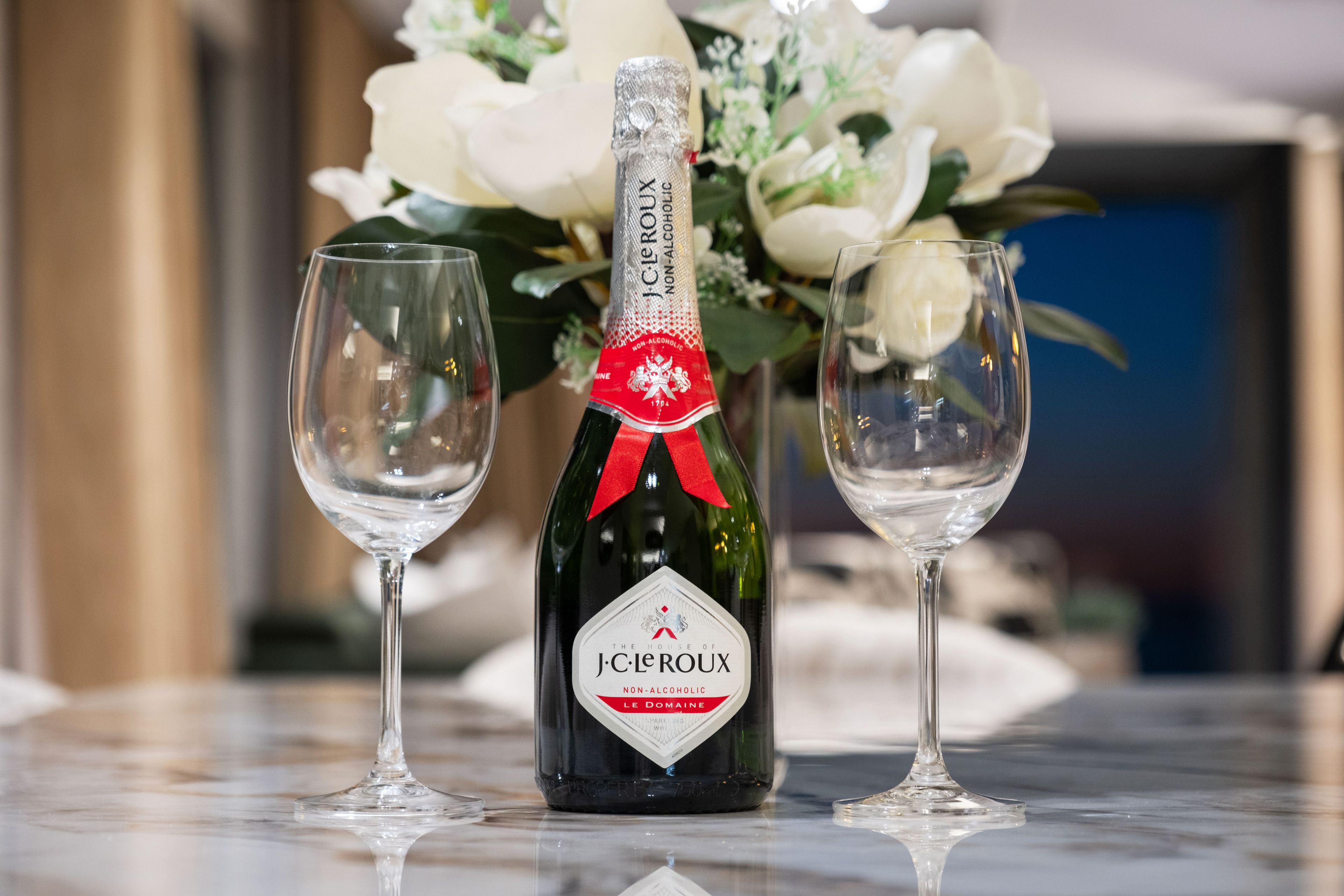 A bottle of J.C. Leroyx Non-Alcoholic wine with a red ribbon, two empty wine glasses, and a floral arrangement in the background.