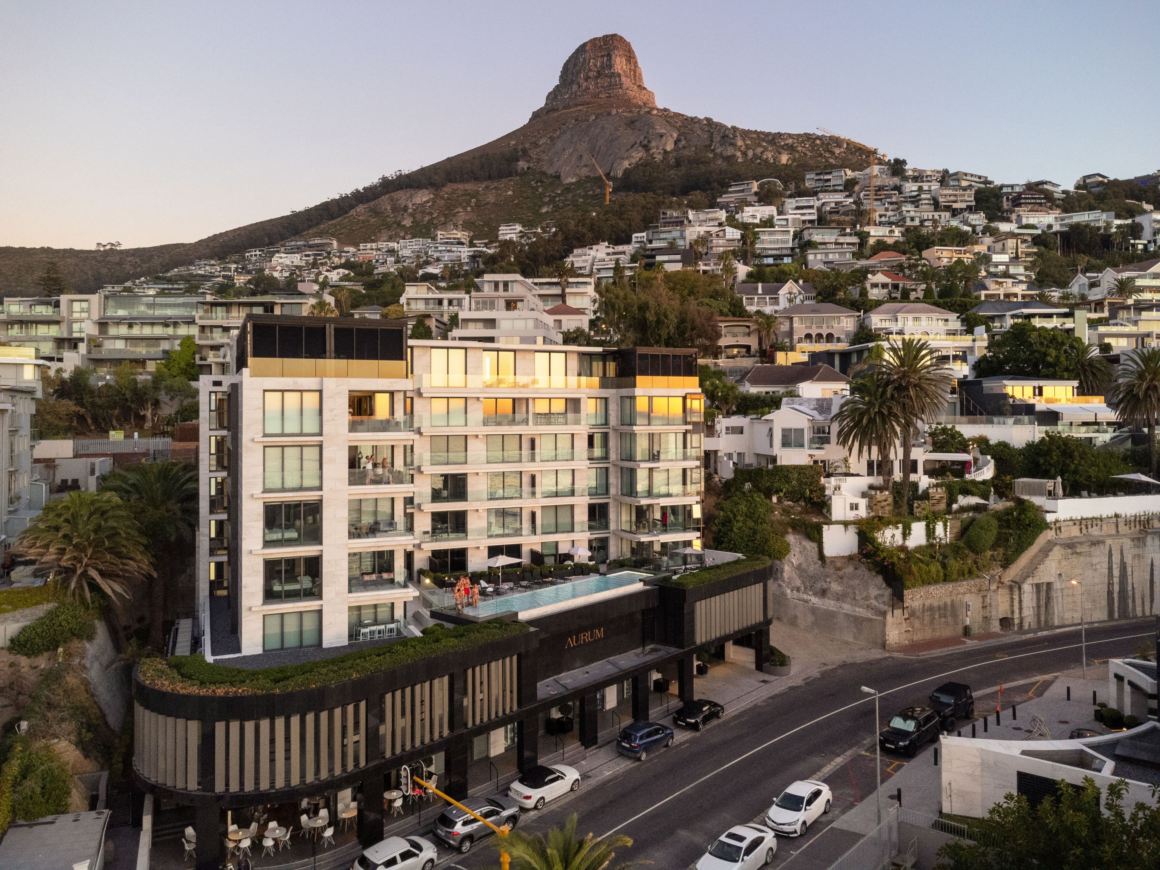 A modern hotel with a rooftop pool, surrounded by a hilly residential area.