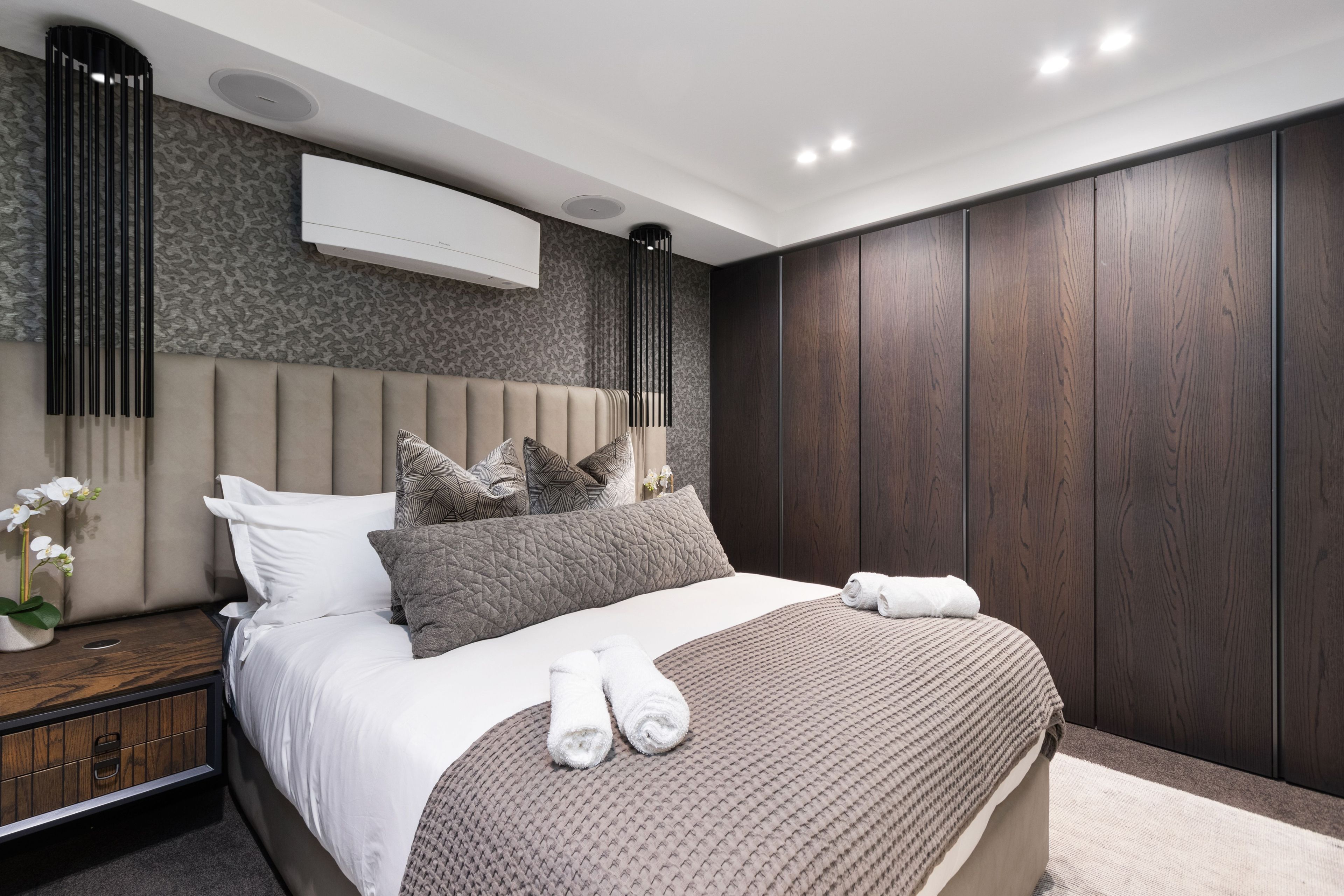 A modern bedroom with a large bed, a nightstand, a wall-mounted air conditioner, and a wooden wardrobe.