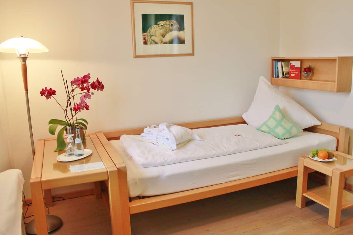 Un lit bien fait, avec du linge de lit blanc, une table de chevet avec une lampe, une petite étagère avec des livres et un vase de fleurs, et un tableau encadré au mur.
