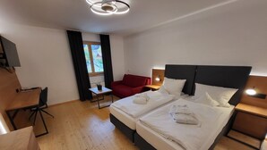 Ein Hotelzimmer mit zwei Betten, einem Schreibtisch, einem roten Sessel, einem kleinen Tisch und einem Fenster mit Vorhängen.