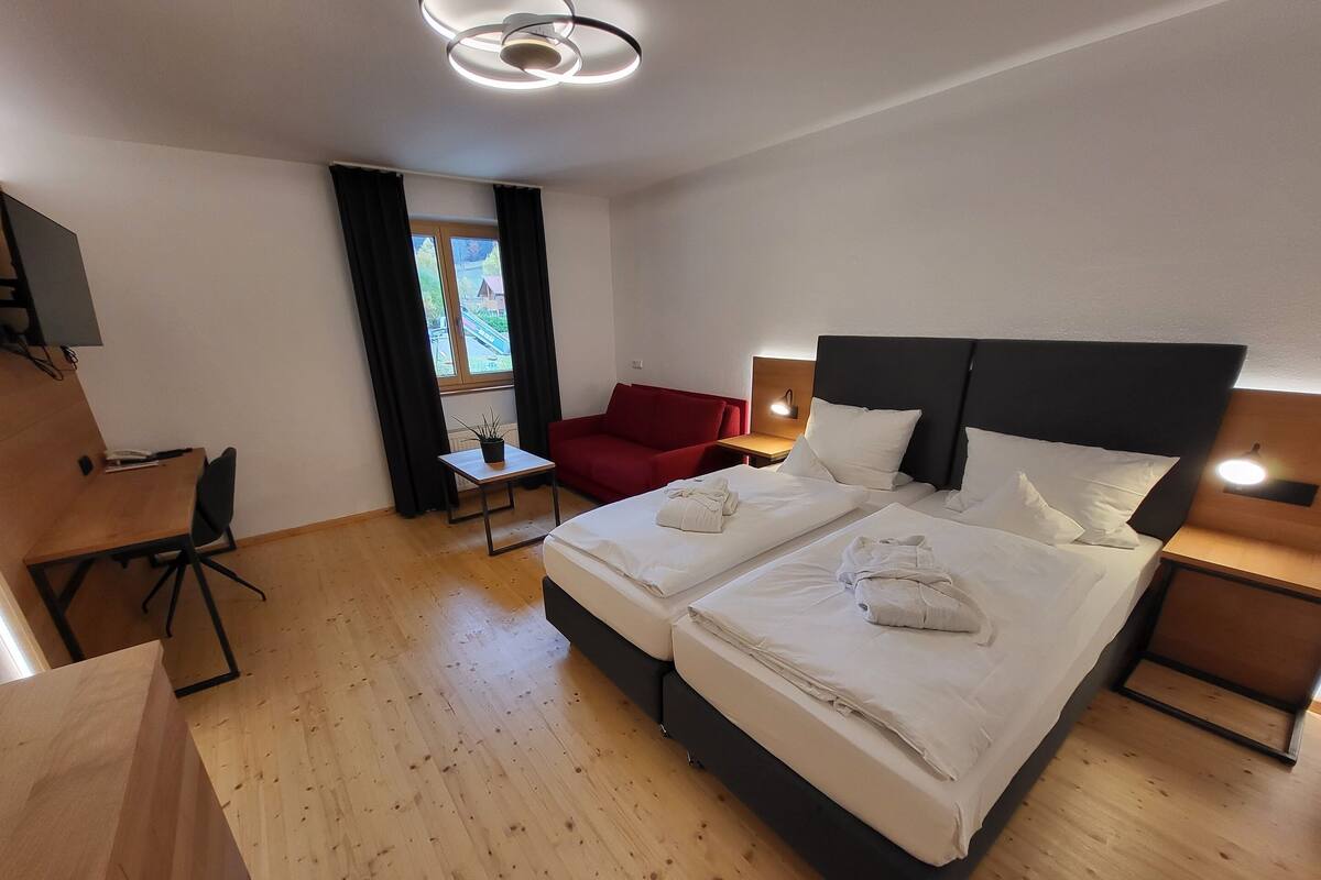 Une chambre d’hôtel avec deux lits, un bureau, une chaise rouge, une petite table et une fenêtre avec des rideaux.