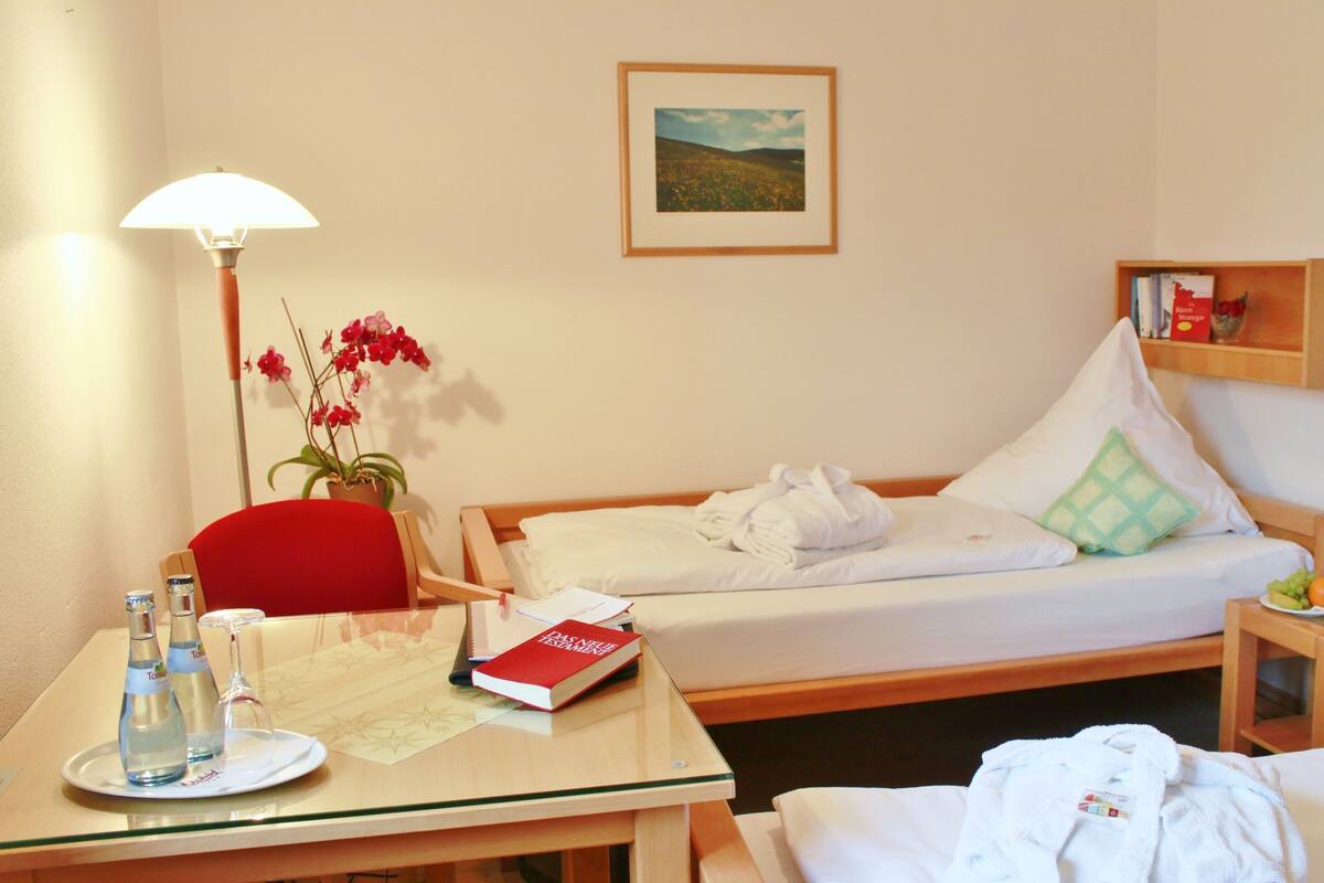 Une chambre d’hôtel avec un lit, un bureau doté d’une lampe, une chaise, une petite table avec un livre et des bouteilles, et une étagère avec des livres et une assiette de fruits.