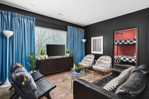 Ein modernes Wohnzimmer mit einer schwarzen Ledersofa, einem Glastisch und einem Fernseher. Im Hintergrund sieht man ein rotes Bett mit karierter Bettwäsche.