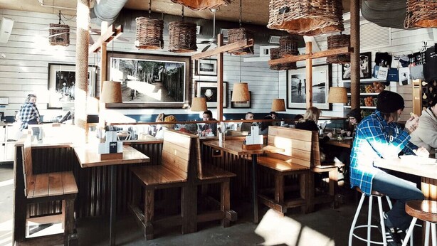 Ein Café mit Holztischen und -stühlen, hängenden Korbwannen und gerahmten Bildern an den Wänden.