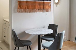 Uma sala de jantar com uma mesa redonda e quatro cadeiras, um armário branco com gavetas e um tecido laranja e branco decorativo pendurado na parede.