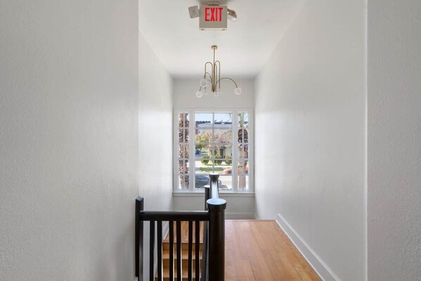 A hallway with a wooden floor, white walls, a black railing, and a window with a view of the outside.