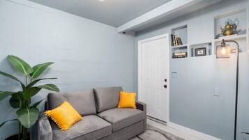 A room with a grey sofa, a potted plant, a white door, and a wall-mounted shelf with books and a teapot.
