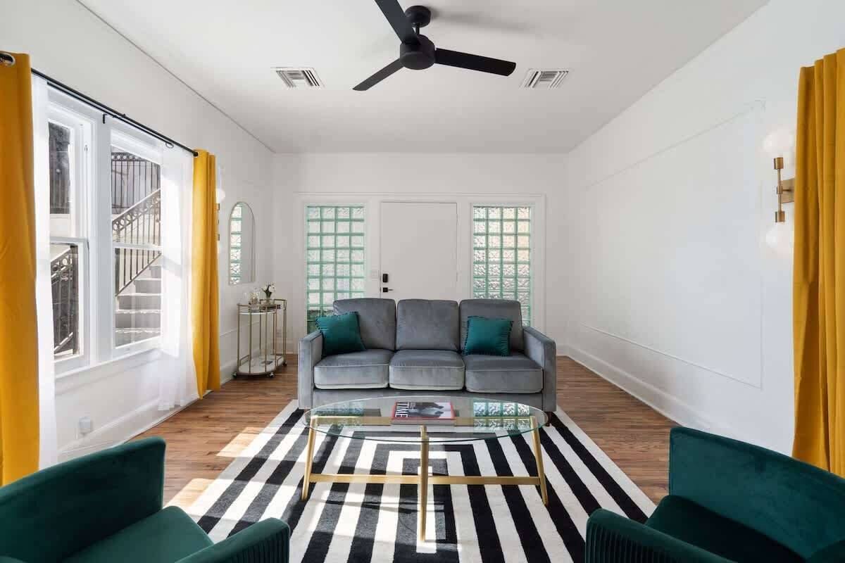Una sala con sofá, mesa de centro y sillas. Se ve un ventilador de techo, una escalera y una puerta con ventanas.