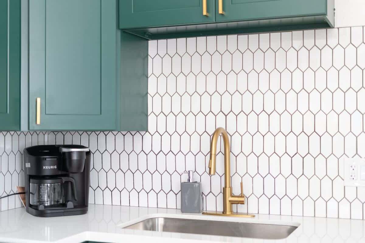 A kitchen with teal cabinets, a white hexagonal tile backsplash, a black Keurig coffee maker, a gold faucet, and a stainless steel sink.