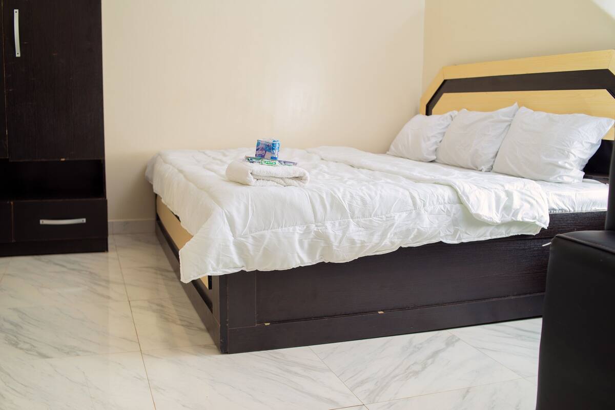 Een netjes opgemaakt bed met witte lakens en een opgevouwen handdoek, een donker houten hoofdbord en een zwart nachtkastje.