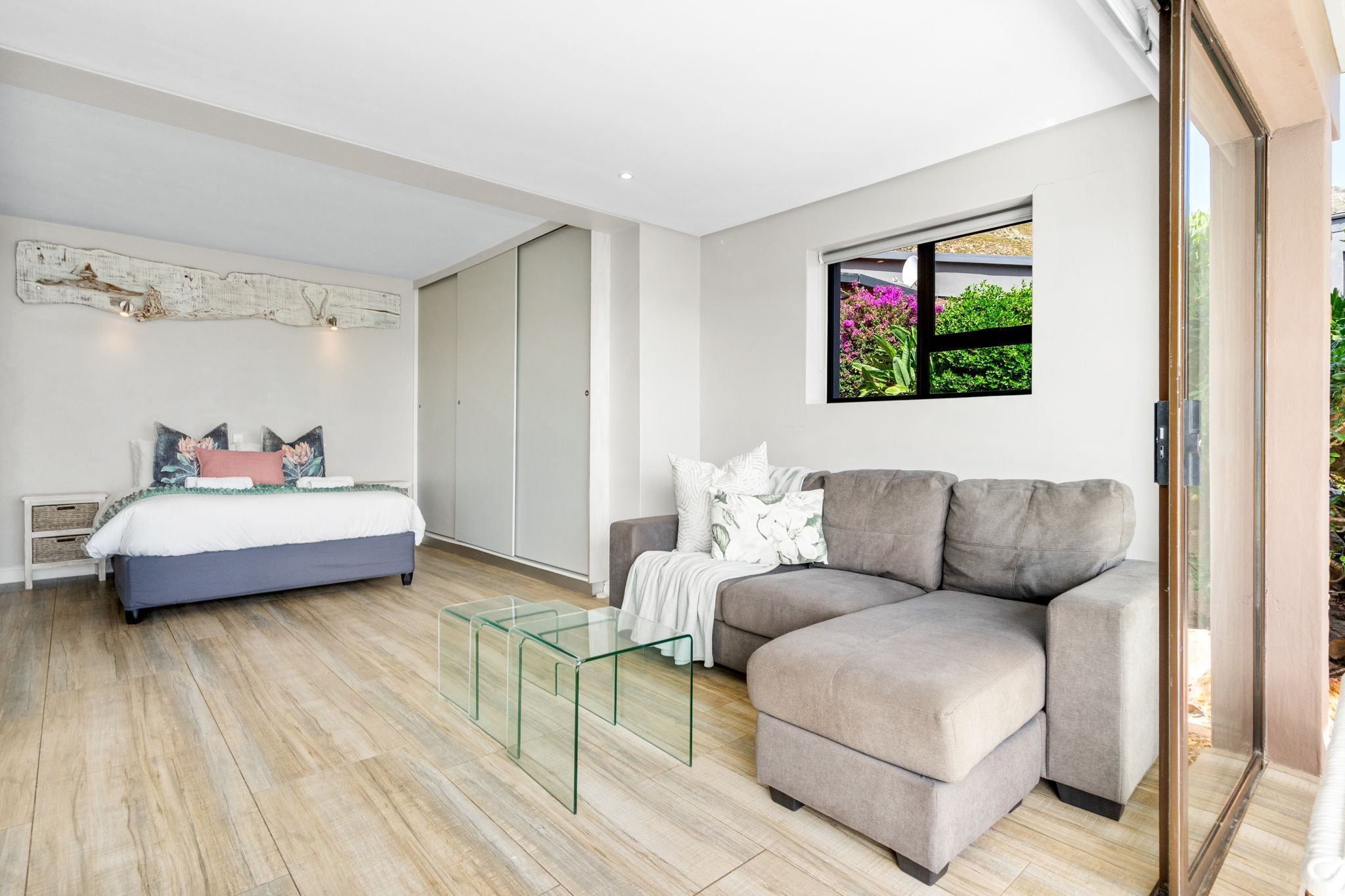 A modern bedroom with a bed, a grey sofa, a glass coffee table, and a window with a view of greenery.