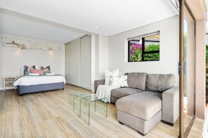 A modern bedroom with a bed, a grey sofa, a glass coffee table, and a window with a view of greenery.